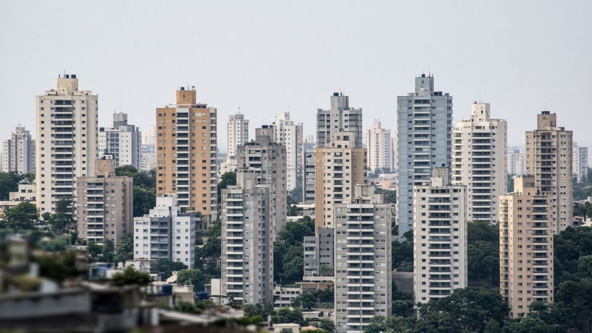 Conjunto denso de torres residenciais de alturas e cores variadas, vistas juntas à distância sobre uma faixa de telhados baixos.